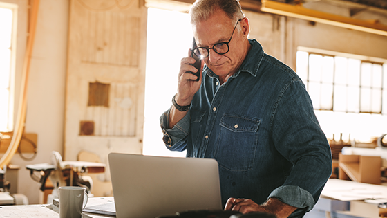 An older man in a warehouse is on his mobile device and laptop.