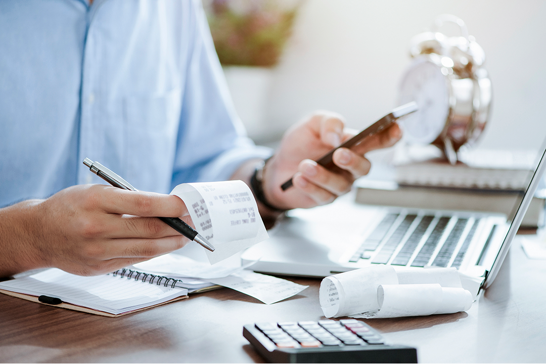 Close up of someone holding receipts with a laptop, mobile device and calculator.