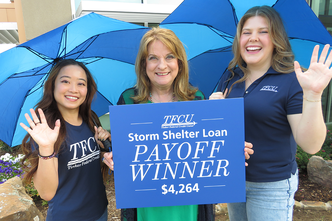 Two TFCU employees and a TFCU member standing together waving at the camera while holding umbrellas and a sign that says storm shelter payoff winner