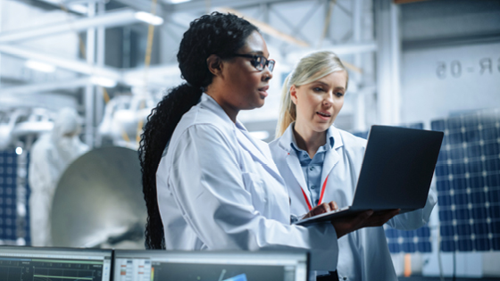 Two women in lab coats in a factory looking at a laptop.