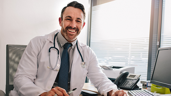 A man who is a doctor is looking and smiling at the camera while working in his office.