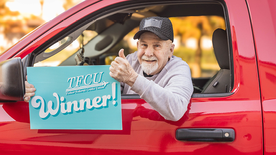 An older man who is a TFCU member is holding a sign while sitting inside a red truck that says winner