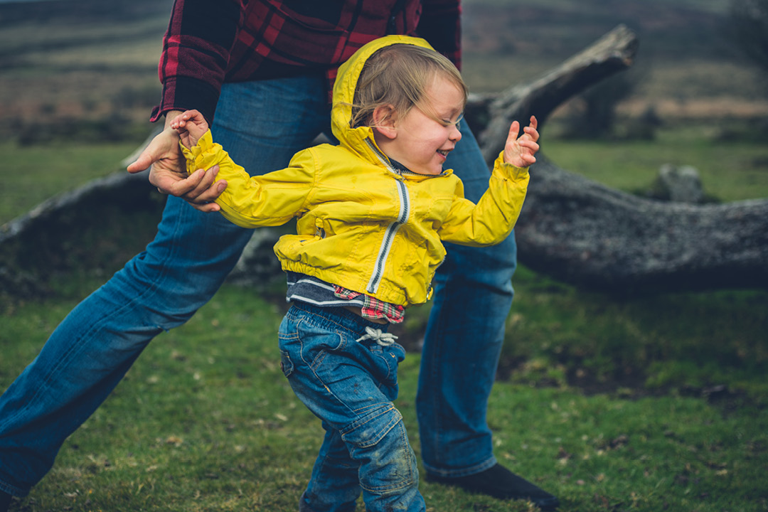 A little boy in a yellow rain coat running in the grass with his dad.