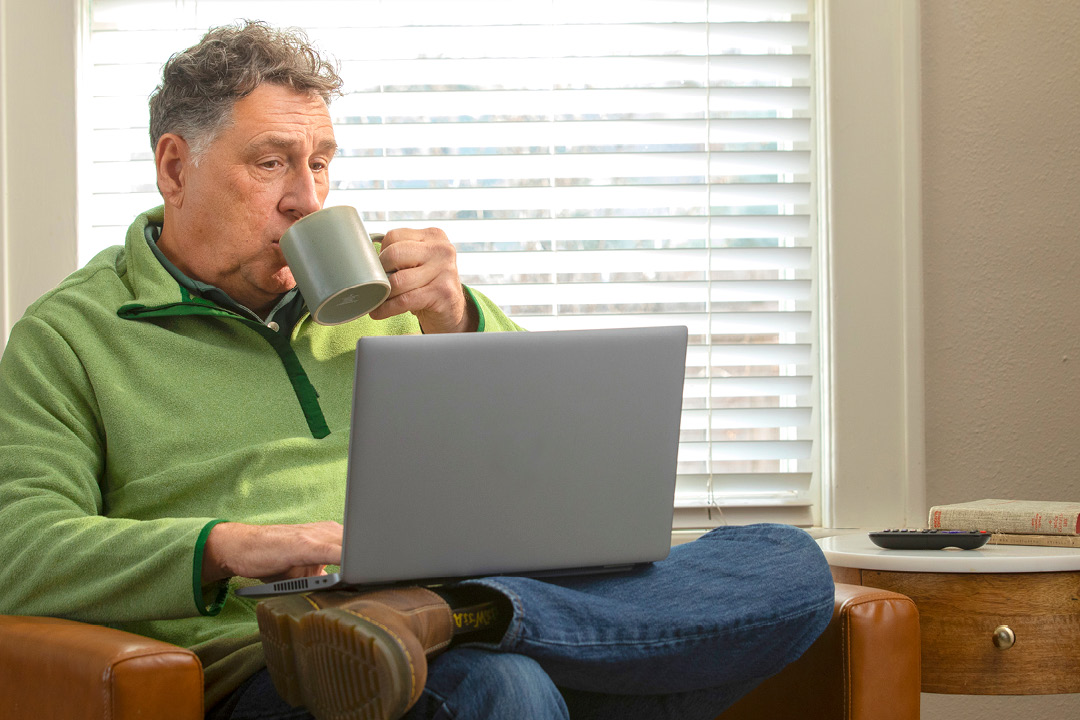 An older man sitting in a chair and drinking out of his mug while on a laptop.