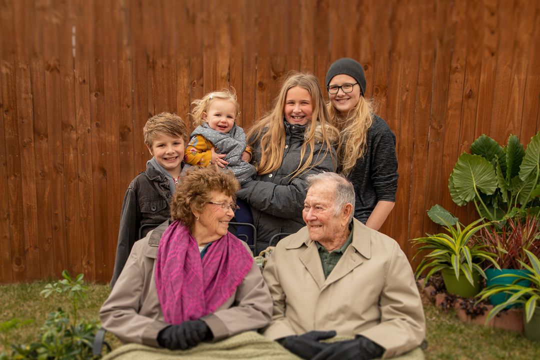 A family group photo with grandparents sitting down and looking at each other and grandchildren in the back