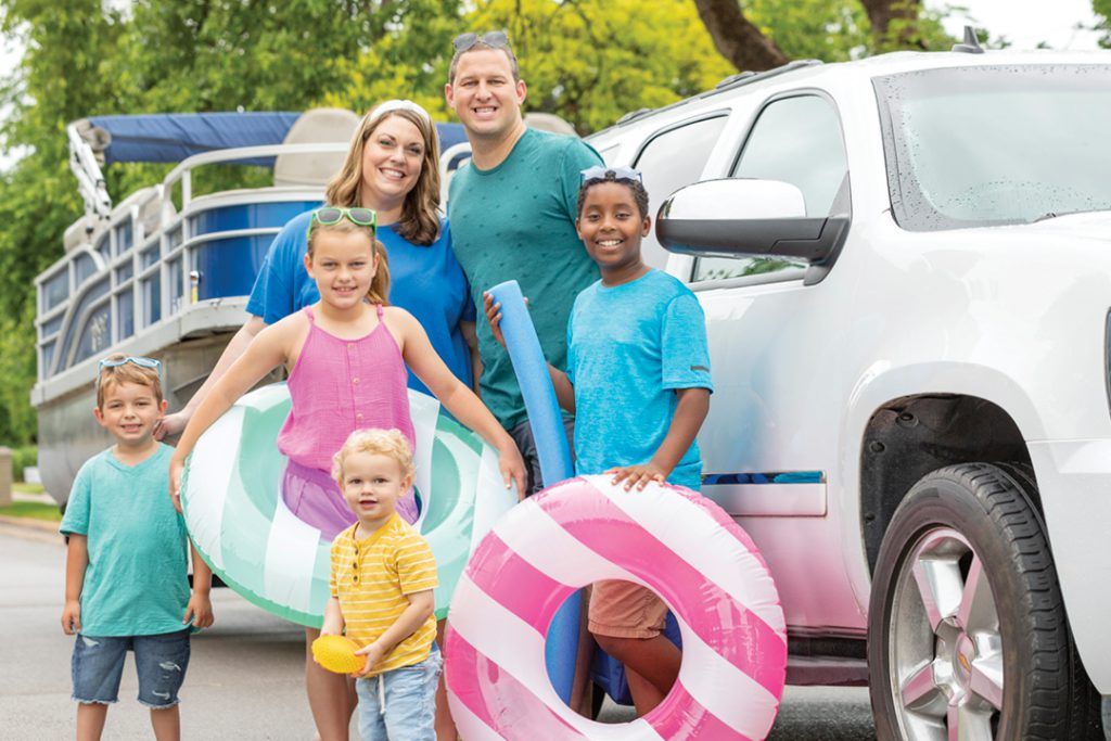 A family of 6 posing and standing in front of an suv and boat